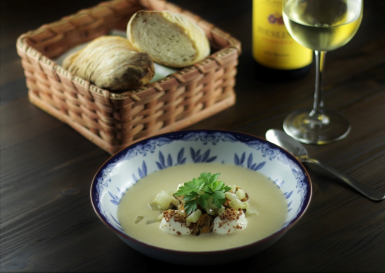 Mussel soup over smoked fresh cheese and sautéed fennel on a table with bread and a glass of wine.